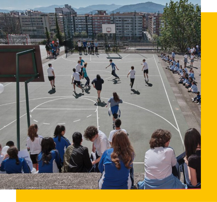Alumnado jugando al baloncesto en el patio del colegio, con compañeros en las gradas.