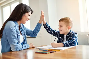 Una mujer y un niño chocan las manos sobre una mesa con material escolar.