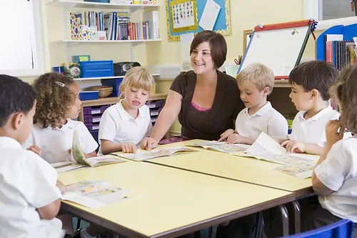 Una docente acompaña a un grupo de niños que trabajan con libros alrededor de una mesa.
