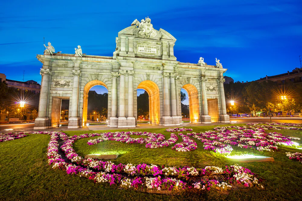 Puerta de Alcalá de Madrid iluminada al anochecer, junto a un jardín de flores